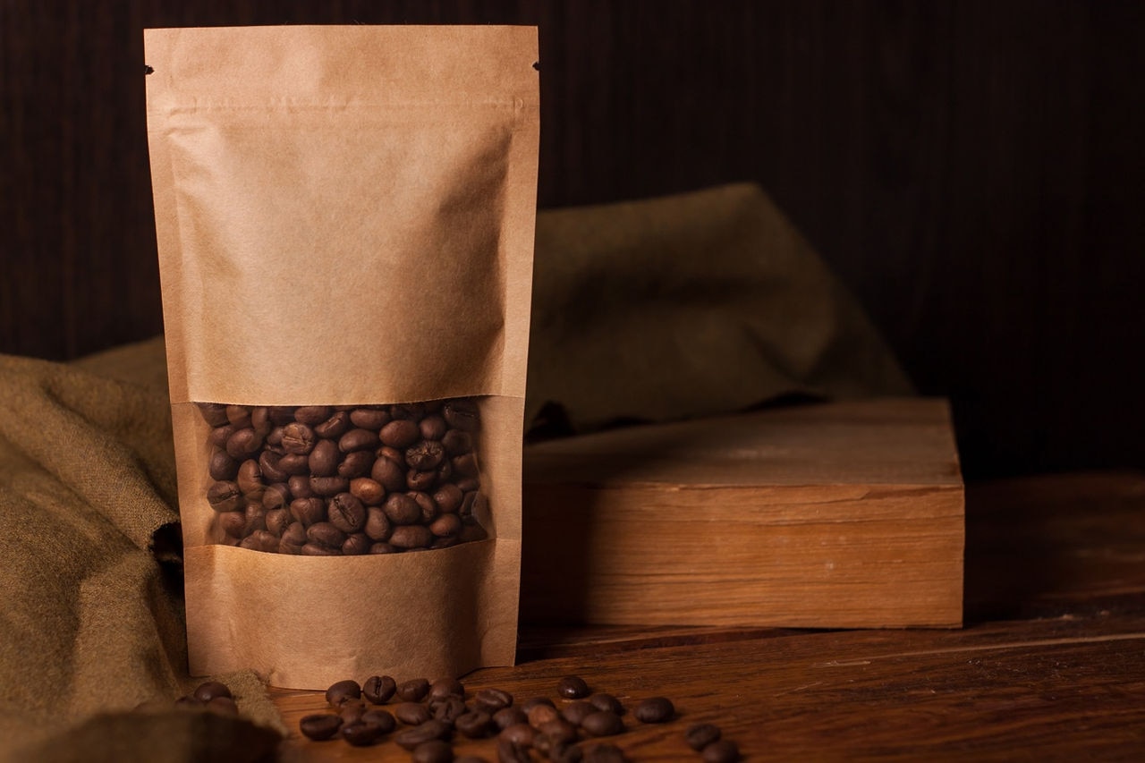 Blank brown kraft paper pouch bag with coffee beans in transparent window on wooden background. Trendy packaging with clasp for tea leaves, herbs, fast food.Mockup for brand name, shopping, hot drinks